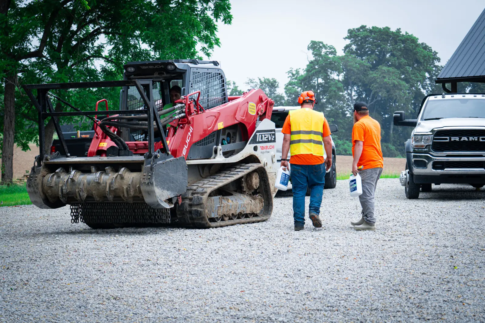 Brushworks crew walking across a gravel yard to a Takeuchi track loader, company RAM truck behind