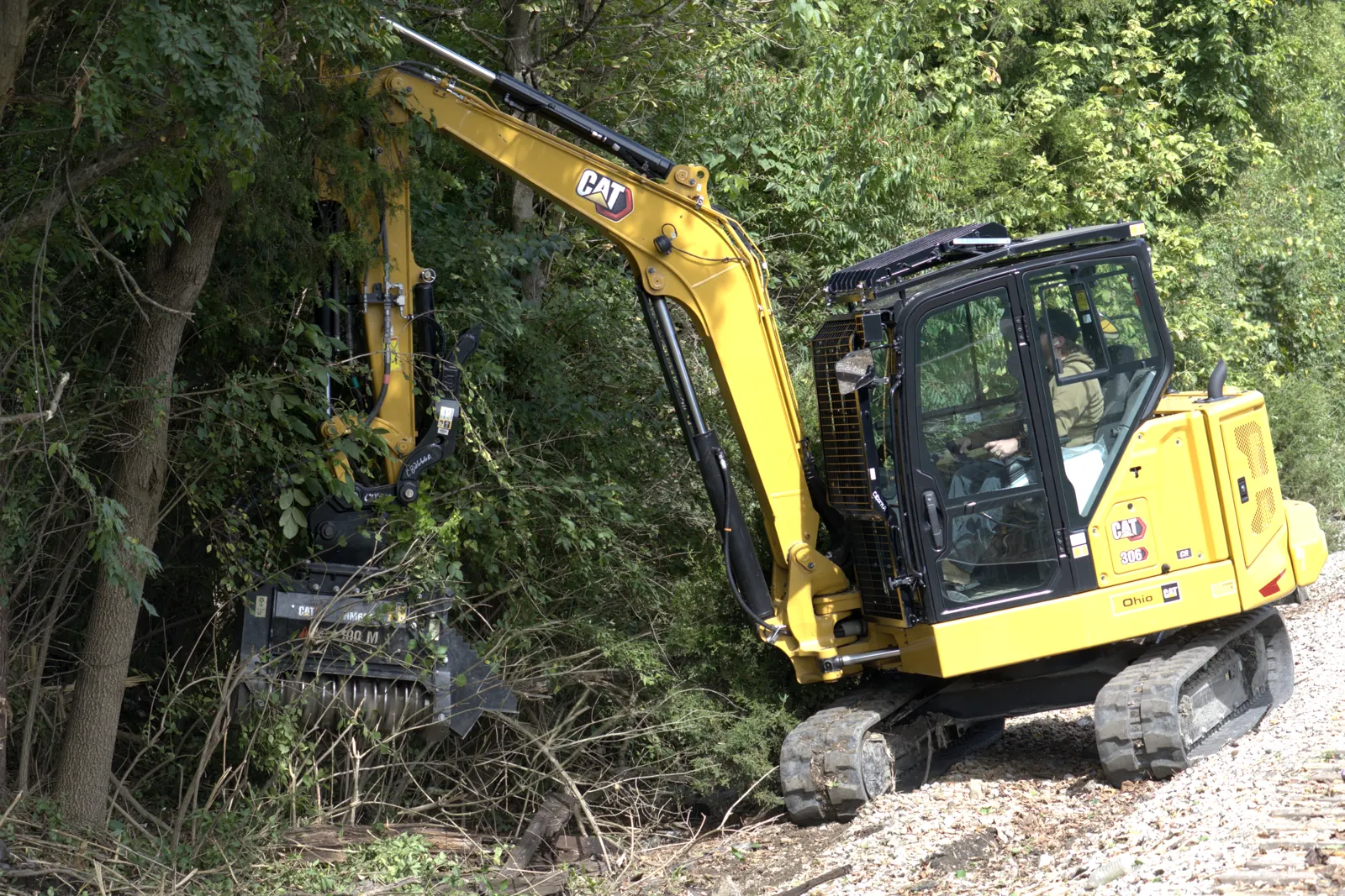 CAT 306 excavator with a mulching head reaching from a gravel edge into a wooded slope