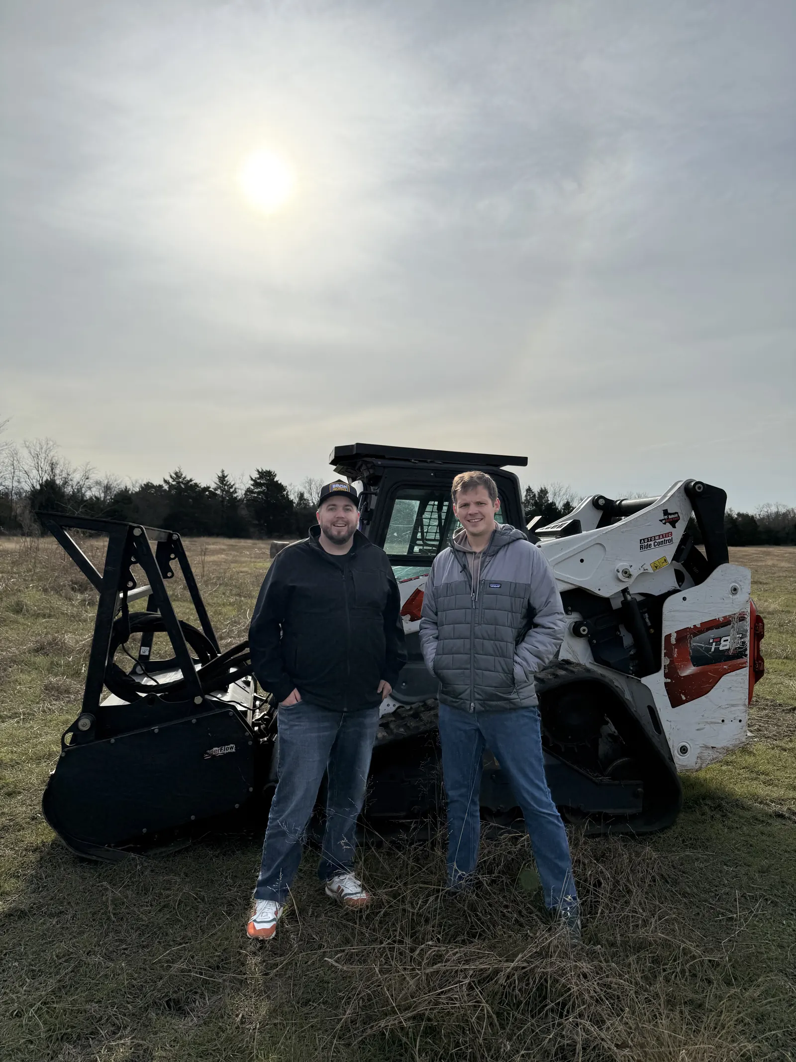 Two Brushworks crew members standing in an open field beside a skid steer
