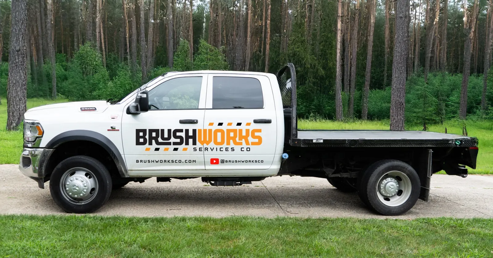 Brushworks branded RAM flatbed work truck parked beside pine trees